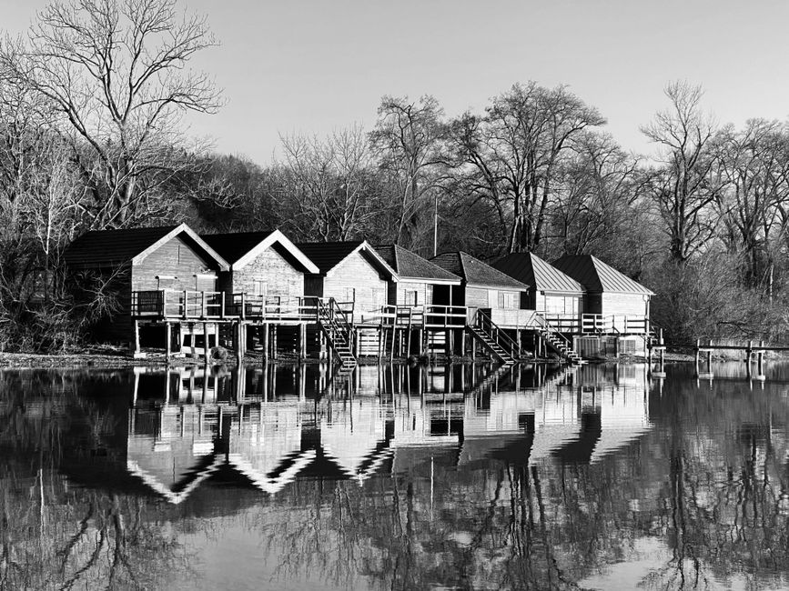 A black and white photograph of a row of wooden lakeside cabins built on stilts over the edge of a calm body of water, with reflections of the structures visible on the water's surface. The cabins hav