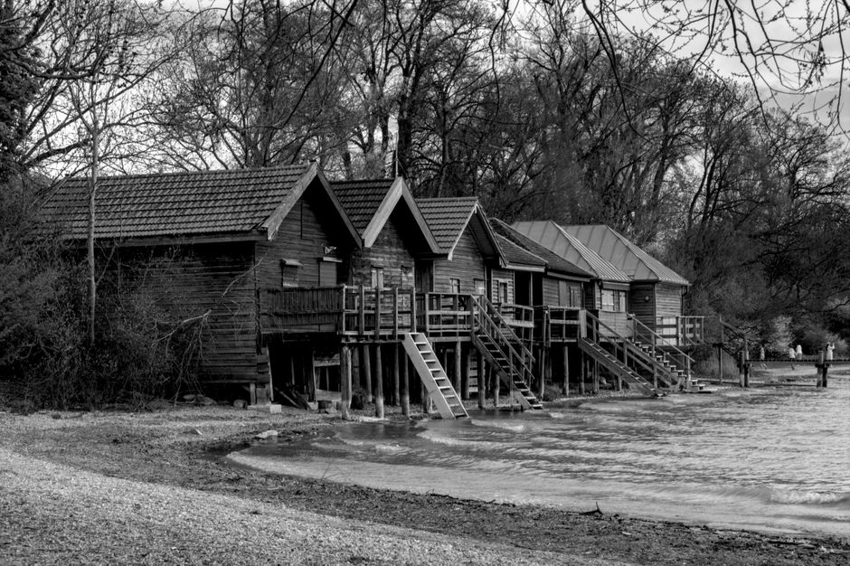 A black and white photograph of a row of wooden lakeside cabins built on stilts over the edge of a calm body of water, with reflections of the structures visible on the water's surface. The cabins hav